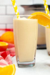 A tall clear glass filled with orange creamsicle Milkshake with a slice of orange in it.