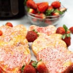 Heart-shaped treats with pink icing and sprinkles grace a white plate, surrounded by fresh strawberries. A bowl of strawberries complements the scene, while a sleek black appliance sits invitingly in the background.