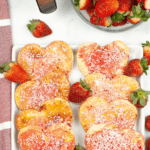 Heart-shaped homemade Pop Tarts with pink icing and white sprinkles rest on a rectangular plate, surrounded by strawberries.