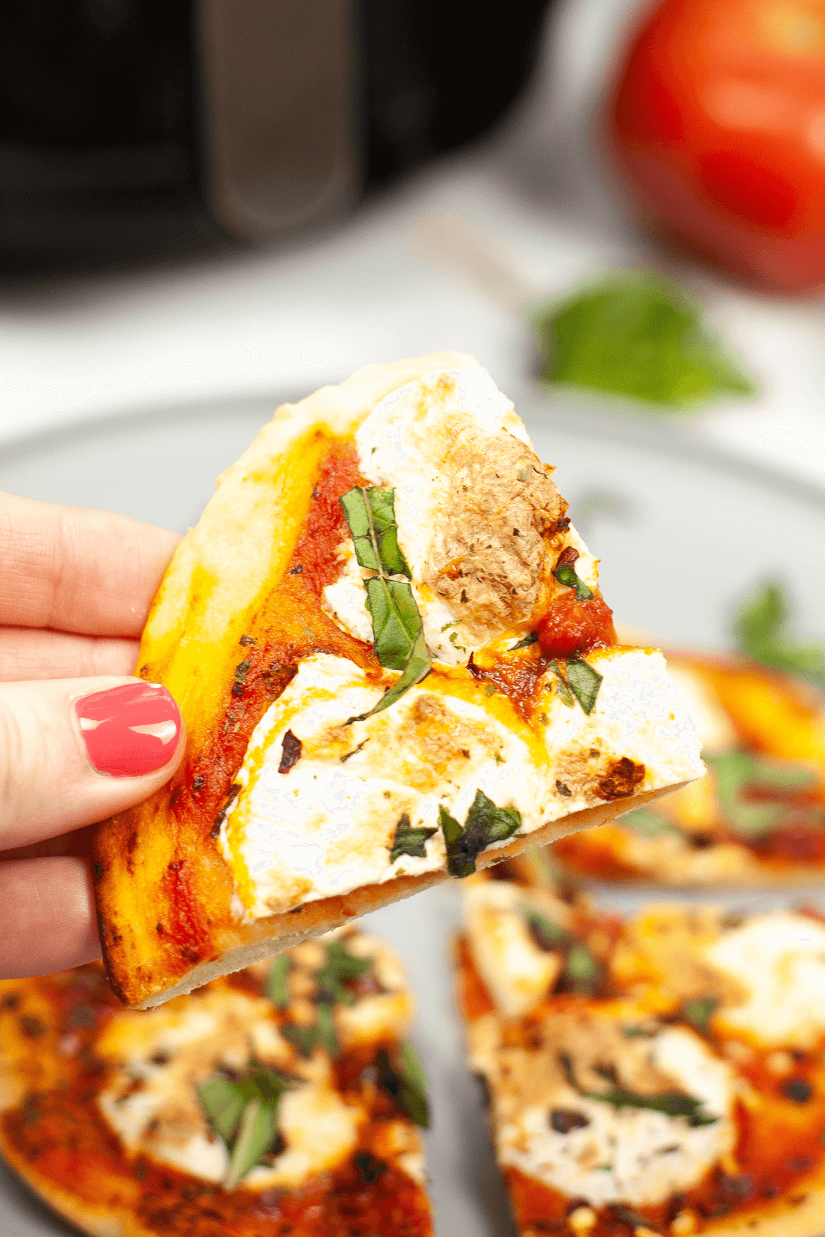 A hand holding a slice of Pizza Margherita topped with cheese, tomato sauce, and fresh basil, with more delightful slices on a plate in the background.