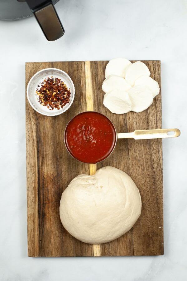 Pizza Margherita ingredients on a wooden board: dough, tomato sauce in a pot, sliced mozzarella, and a dish of red pepper flakes.