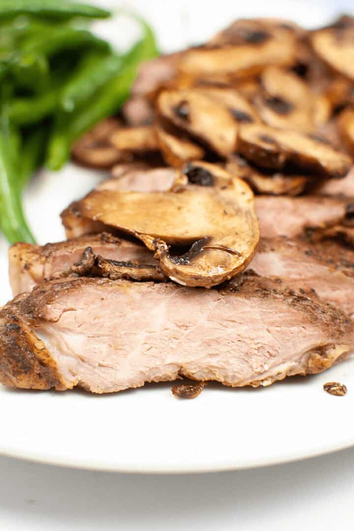 Sliced air fryer ribeye steak topped with sautéed mushrooms, served alongside green beans on a pristine white plate.