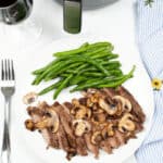 A plate of air fryer ribeye steak slices, accompanied by mushrooms and green beans, sits invitingly next to a fork and a glass of red wine on the table.