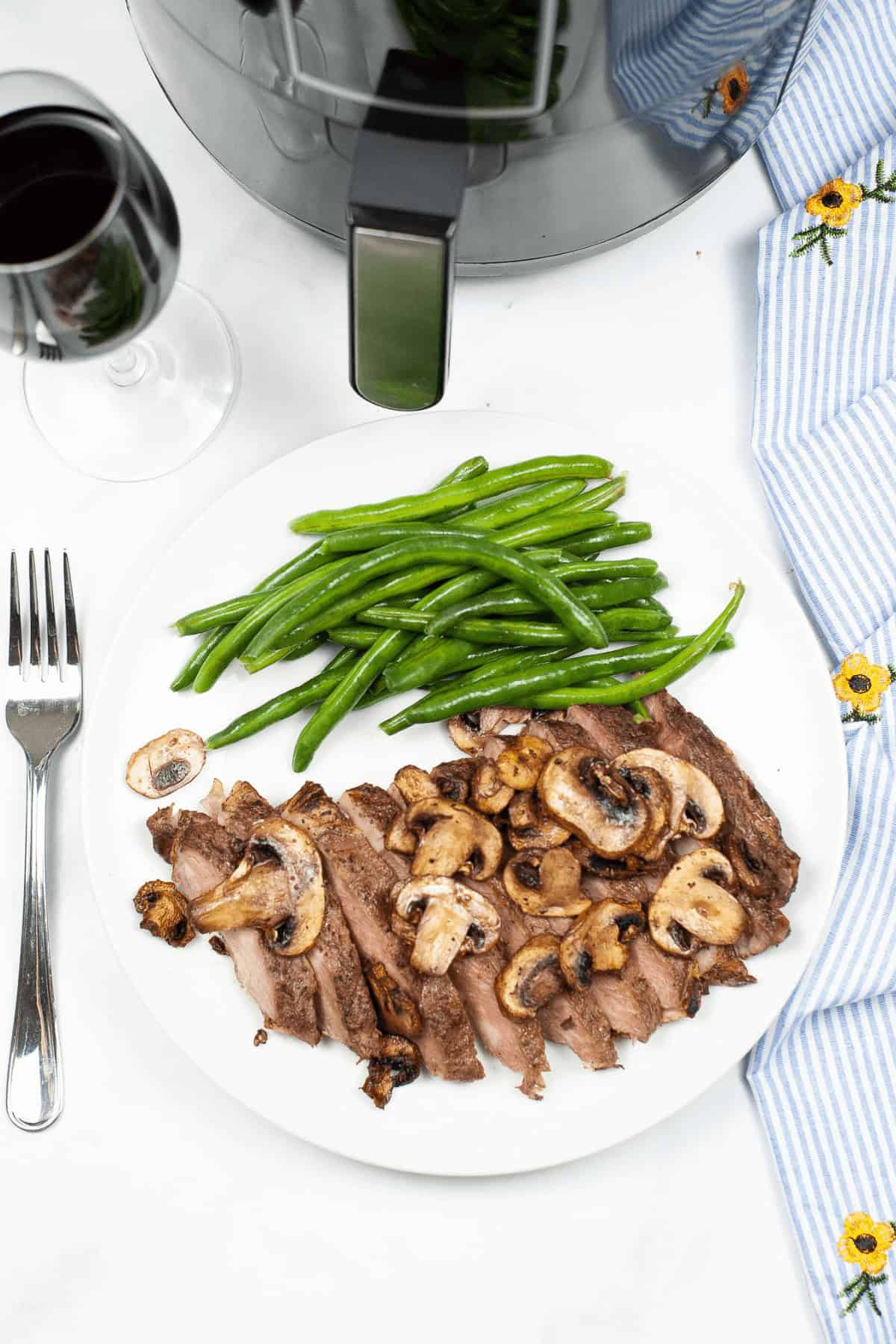 A plate of air fryer ribeye steak slices, accompanied by mushrooms and green beans, sits invitingly next to a fork and a glass of red wine on the table.