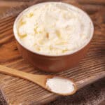 A wooden bowl filled with homemade whipped butter sits on a wooden board, accompanied by a small wooden spoon with a dab of creamy butter on it.