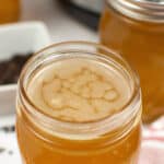 Close-up of a mason jar filled with light brown Instant Pot chicken bone broth, next to another jar. A white dish with dark spices is partially visible in the background.