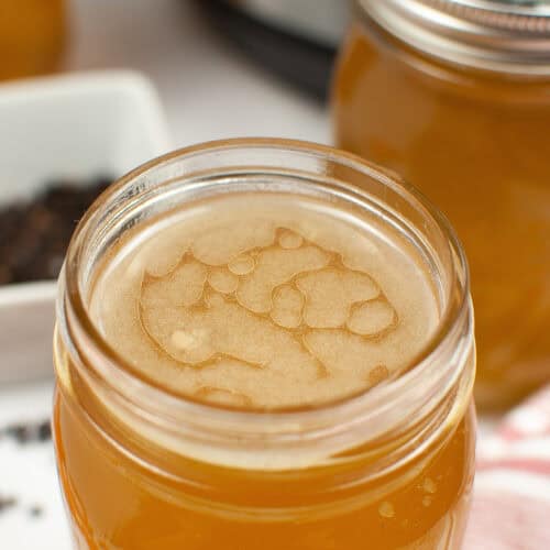 Close-up of a mason jar filled with light brown Instant Pot chicken bone broth, next to another jar. A white dish with dark spices is partially visible in the background.