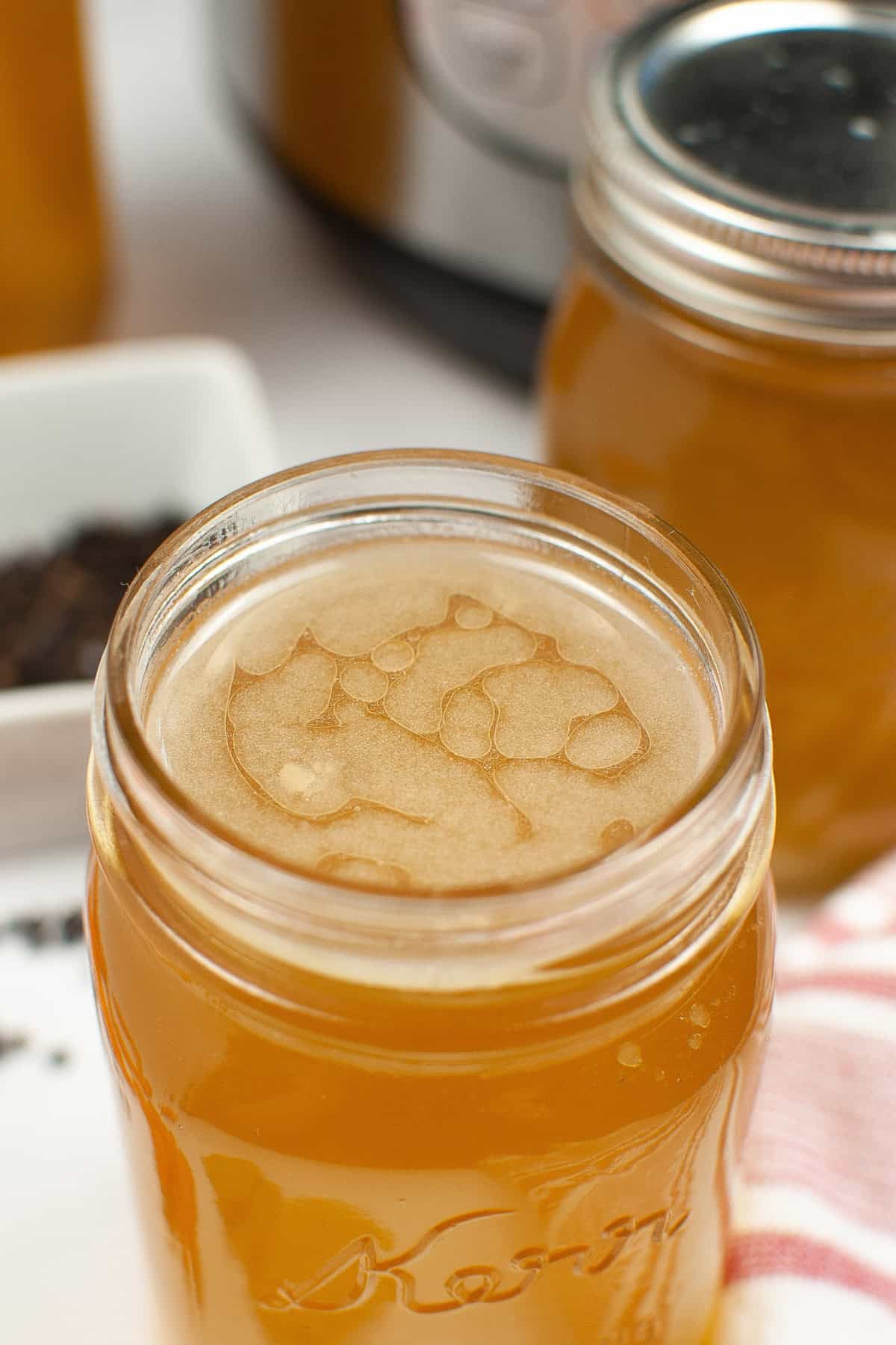 Close-up of a mason jar filled with light brown Instant Pot chicken bone broth, next to another jar. A white dish with dark spices is partially visible in the background.
