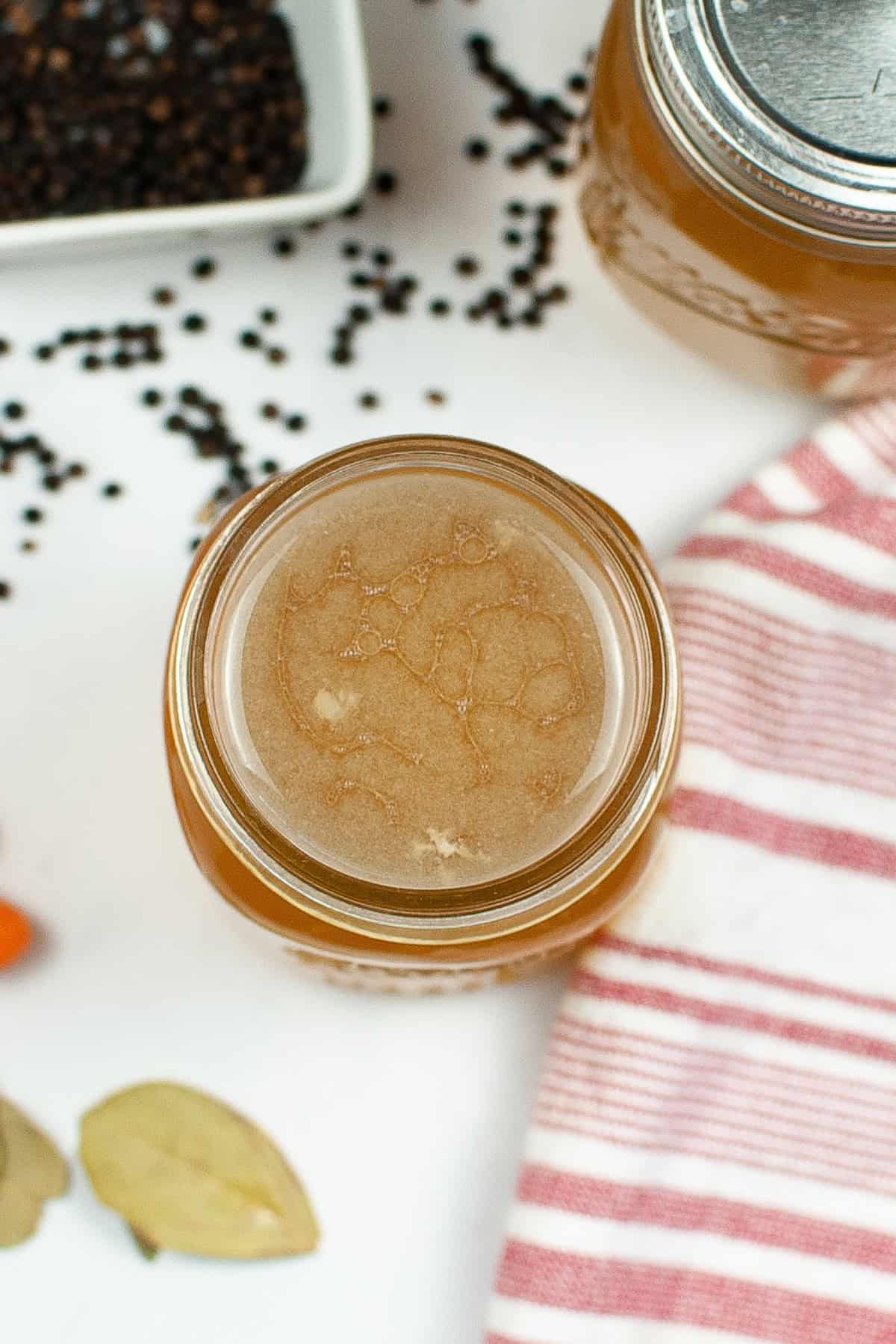 Top view of a jar filled with liquid, surrounded by a striped cloth, black peppercorns, and bay leaves, suggesting a homemade concoction preparation.