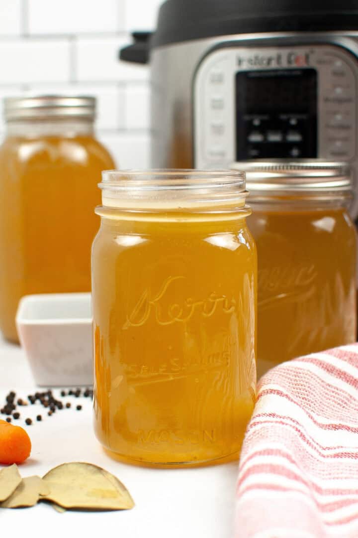 Mason jars brimming with Instant Pot chicken bone broth sit proudly on the counter. Nearby, a dish towel flutters beside peppercorns, garlic, turmeric, and bay leaves—each ingredient having played its part in transforming simple elements into a nourishing elixir.