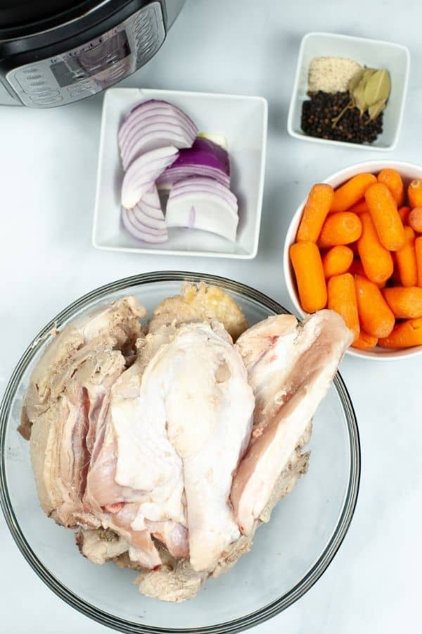 Ingredients for cooking: a bowl of raw chicken, a bowl of baby carrots, sliced red onion, and a blend of spices with bay leaves ready near an electric cooker.