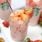 A mason jar filled with a pink strawberry drink, ice, and slices of fresh strawberries. Two Starbucks Pink Drinks in the background, and fresh strawberries are scattered around.