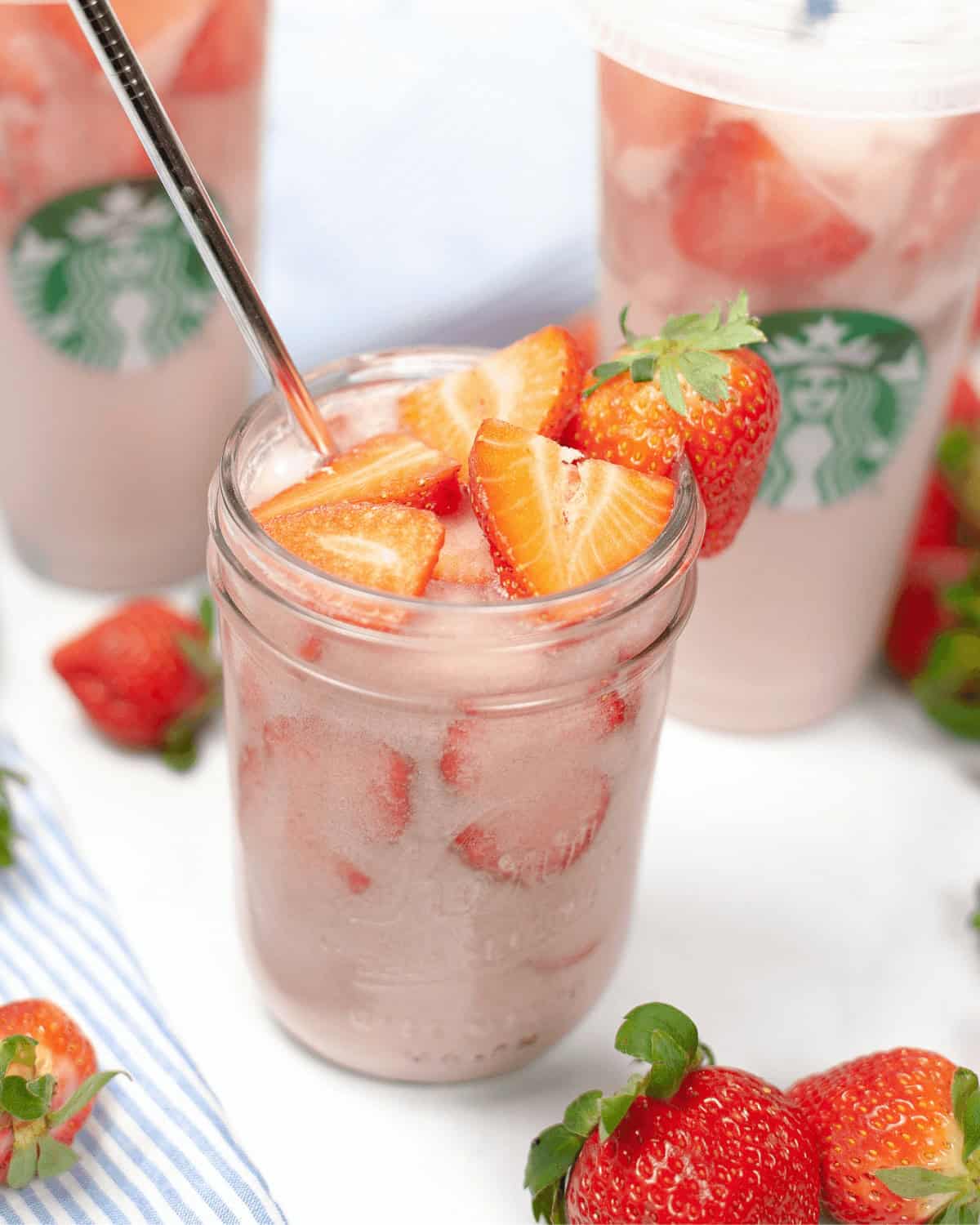 A mason jar filled with a pink strawberry drink, ice, and slices of fresh strawberries. Two Starbucks Pink Drinks in the background, and fresh strawberries are scattered around.