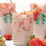 Three strawberry beverages with ice; two Starbucks Pink Drinks in branded cups in the background and one in a glass jar with a metal straw in the foreground, surrounded by fresh strawberries.