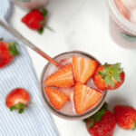 A Starbucks Pink Drink in a glass with ice and sliced strawberries, alongside a striped cloth and whole strawberries.