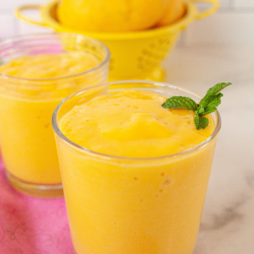 two glasses of mango smoothie with a colander filled with mango in the background.