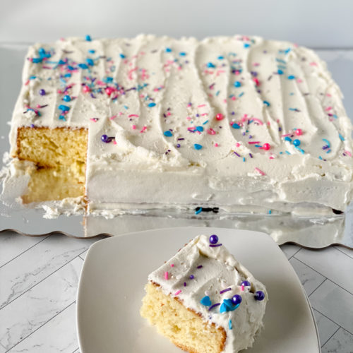 Slice of Homemade Vanilla Cake on a white plate alongside a full cake.