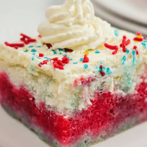 A slice of 4th of July Poke Cake with red, white, and blue layers topped with whipped cream and multicolored sprinkles on a white plate.