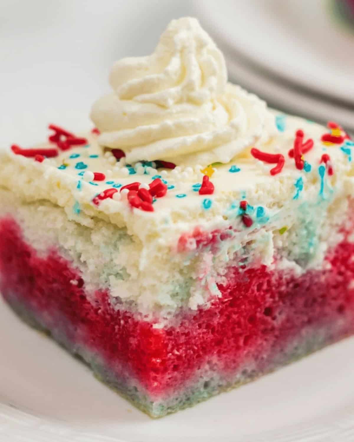 A slice of 4th of July Poke Cake with red, white, and blue layers topped with whipped cream and multicolored sprinkles on a white plate.