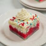 A slice of 4th of July Poke Cake features tri-colored layers topped with white frosting, colorful sprinkles, and a swirl of whipped cream on a white plate.