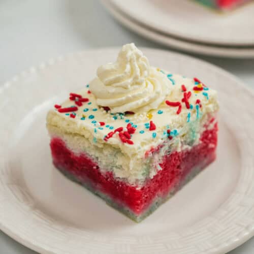 A slice of 4th of July Poke Cake features tri-colored layers topped with white frosting, colorful sprinkles, and a swirl of whipped cream on a white plate.