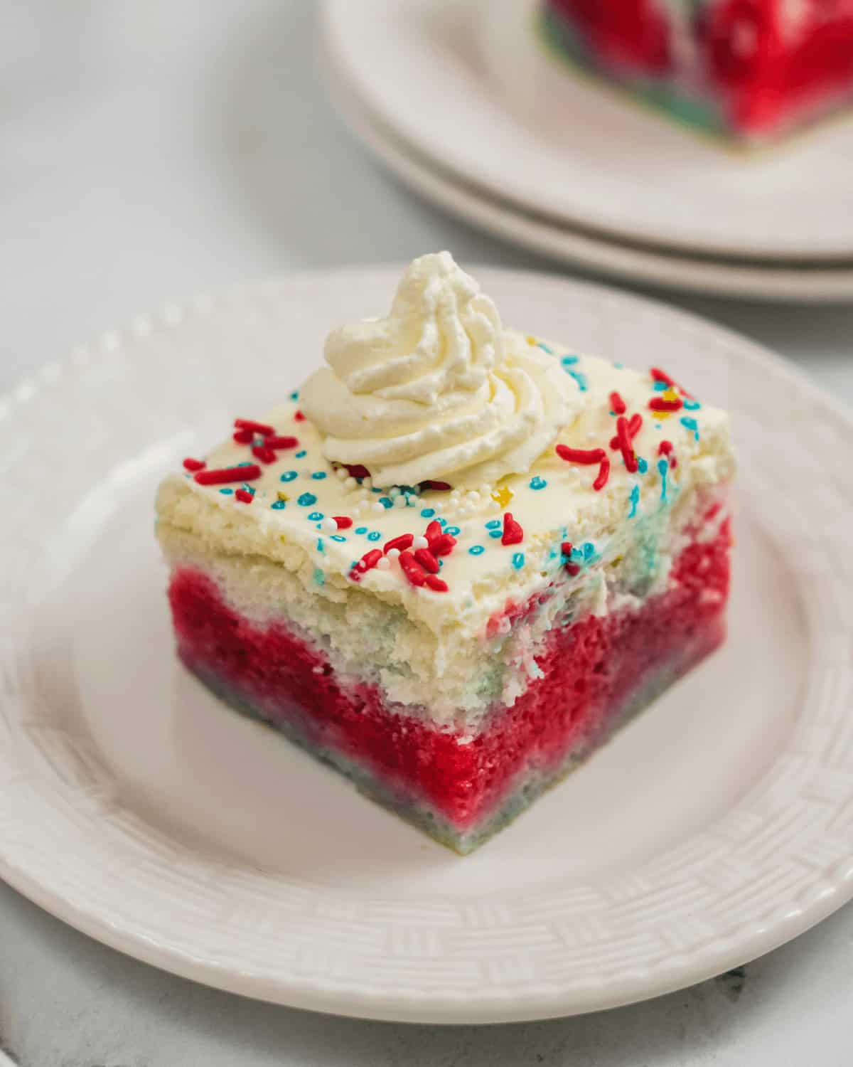 A slice of 4th of July Poke Cake features tri-colored layers topped with white frosting, colorful sprinkles, and a swirl of whipped cream on a white plate.