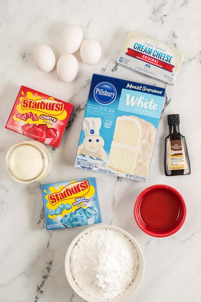 Ingredients on a marble surface: four eggs, cream cheese, white cake mix, cherry Starburst candy, blue raspberry Starburst candy, vanilla extract, heavy cream, powdered sugar, and a small red bowl — perfect for making a 4th of July Poke Cake.