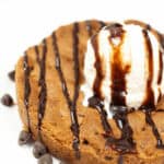 A classic pizookie: a warm chocolate chip cookie topped with a scoop of vanilla ice cream and drizzled with rich chocolate syrup, all served on a pristine white plate.
