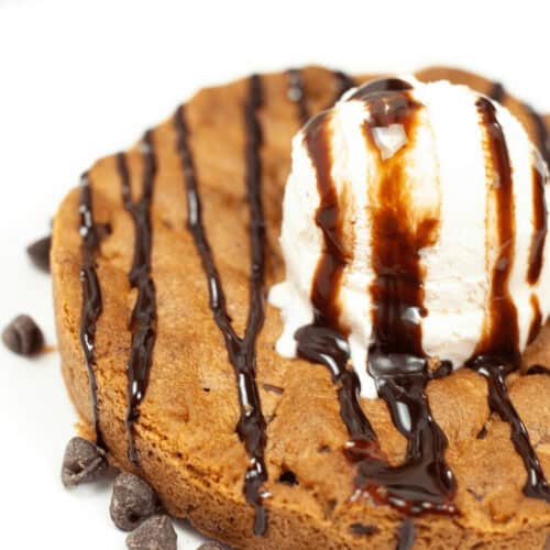 A classic pizookie: a warm chocolate chip cookie topped with a scoop of vanilla ice cream and drizzled with rich chocolate syrup, all served on a pristine white plate.