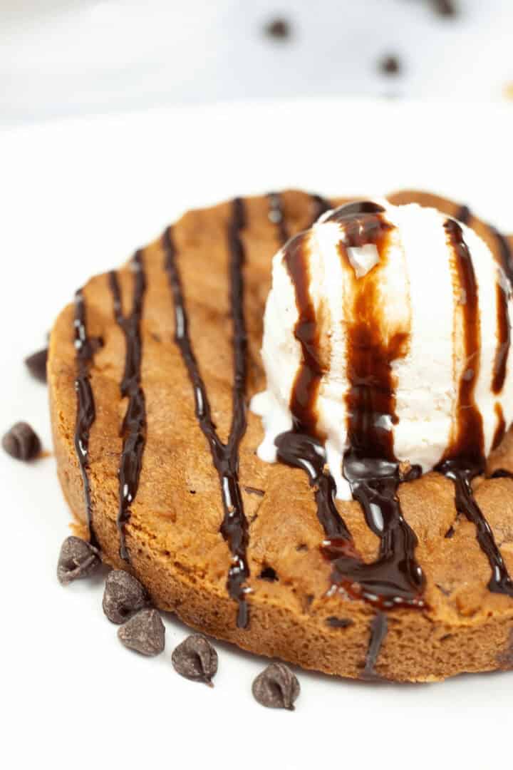 A classic pizookie: a warm chocolate chip cookie topped with a scoop of vanilla ice cream and drizzled with rich chocolate syrup, all served on a pristine white plate.