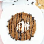 A decadent cookie—a large chocolate chip cookie topped with a creamy scoop of vanilla ice cream—drizzled with rich chocolate sauce, beautifully displayed on a white plate, complete with scattered chocolate chips for extra indulgence.