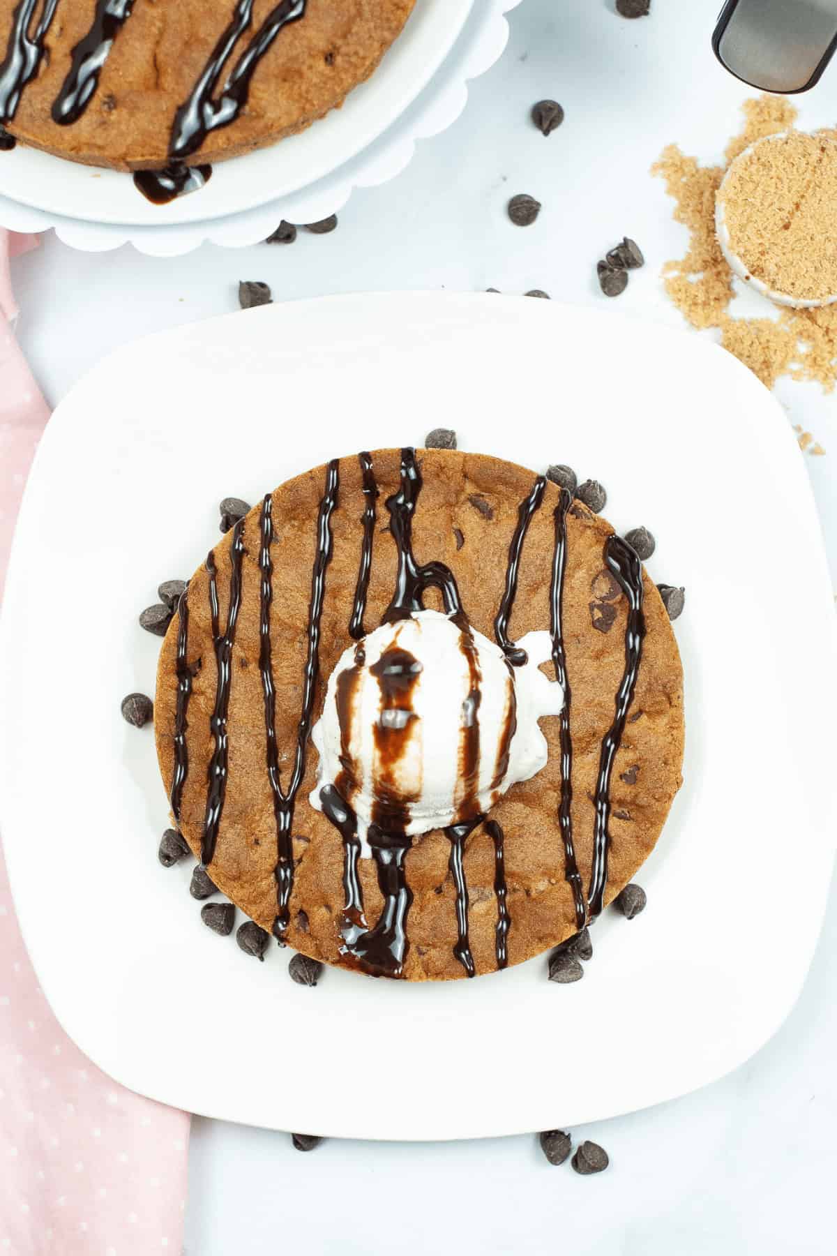 A decadent cookie—a large chocolate chip cookie topped with a creamy scoop of vanilla ice cream—drizzled with rich chocolate sauce, beautifully displayed on a white plate, complete with scattered chocolate chips for extra indulgence.