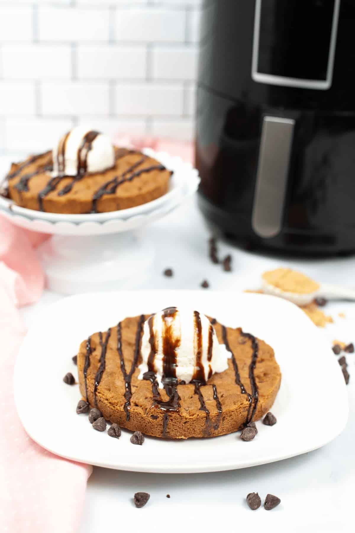 Two chocolate chip cookie cakes, reminiscent of a delightful pizookie, are topped with ice cream and chocolate drizzle on plates, with an air fryer and some chocolate chips in the background.
