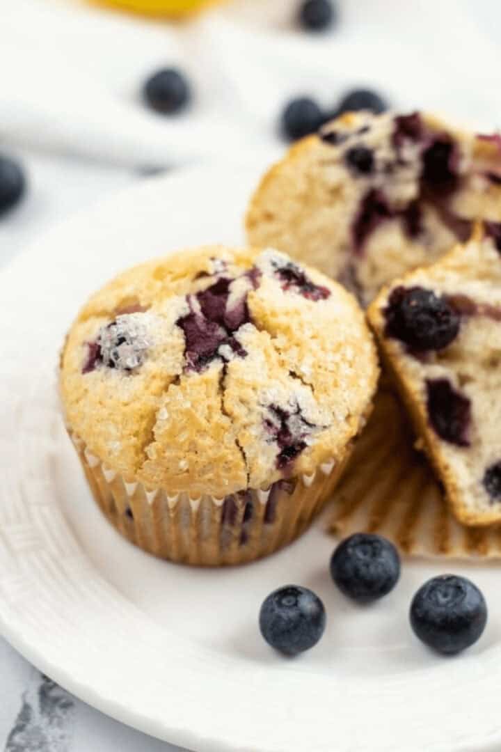 A blueberry lemon muffin with a cracked top sits on a white plate, accompanied by a halved muffin and scattered blueberries.