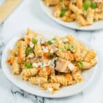 A plate of the finished dish of chicken, crumbled feta cheese, chopped green onions, and a creamy dressing rests on a marble surface. Another plate and wooden utensil are blurred in the background.