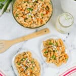 A glass bowl and two plates of chicken pasta salad with rotini, peas, diced vegetables, and cheese sit on the marble surface. Accompanied by a wooden spatula, a jar with an ice drink, and a striped napkin.