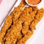 A white rectangular plate with crunchy chicken tenders, reminiscent of classic finger steaks, alongside a small bowl of dipping sauce. A red and white checkered cloth is partially visible in the background.