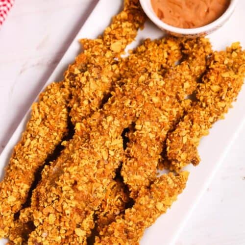 A white rectangular plate with crunchy chicken tenders, reminiscent of classic finger steaks, alongside a small bowl of dipping sauce. A red and white checkered cloth is partially visible in the background.