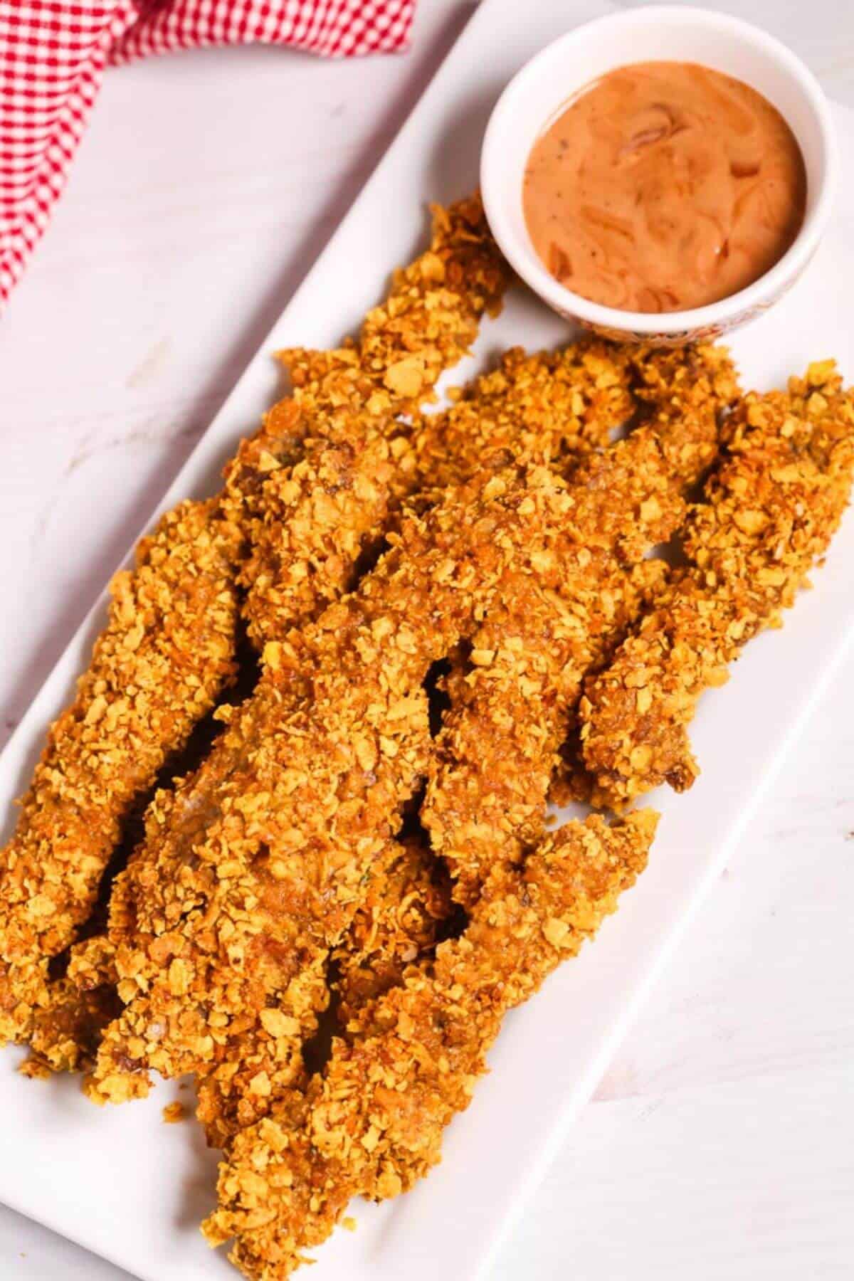 A white rectangular plate with crunchy chicken tenders, reminiscent of classic finger steaks, alongside a small bowl of dipping sauce. A red and white checkered cloth is partially visible in the background.