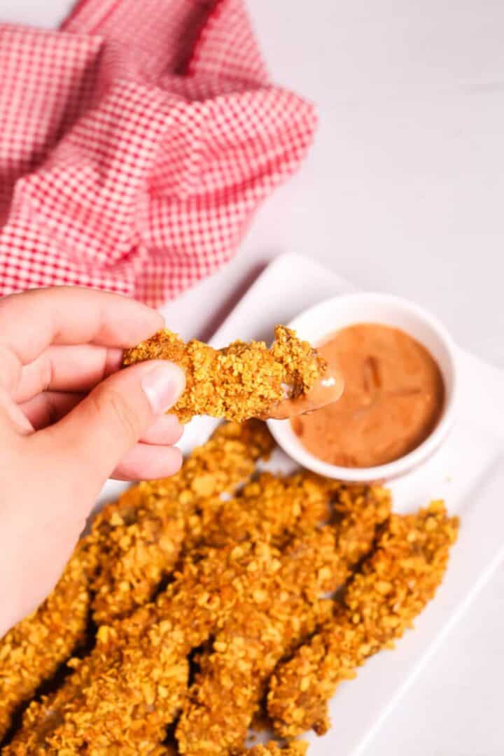 A hand holds a finger steak dipped in sauce, with more crispy chicken strips on a white plate beside a bowl of dipping sauce. A red and white checkered cloth adds a cozy background touch.