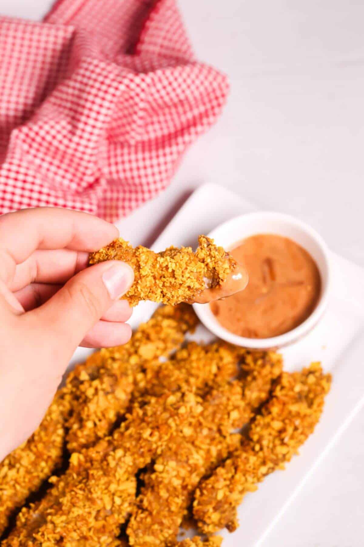 A hand holds a finger steak dipped in sauce, with more crispy chicken strips on a white plate beside a bowl of dipping sauce. A red and white checkered cloth adds a cozy background touch.