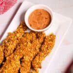 Crispy breaded finger steaks are artfully arranged on a rectangular white plate, accompanied by a small bowl of dipping sauce. In the background, a red checkered cloth adds a touch of classic charm to this delightful setup.