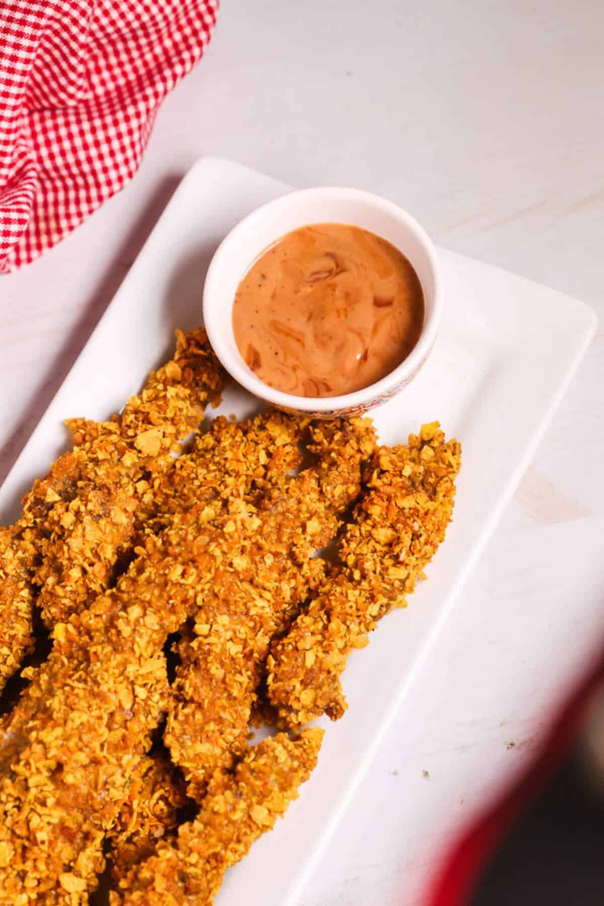 Crispy breaded finger steaks are artfully arranged on a rectangular white plate, accompanied by a small bowl of dipping sauce. In the background, a red checkered cloth adds a touch of classic charm to this delightful setup.