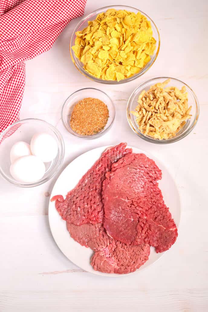 Ingredients for arranged on a table: raw beef slices on a plate, eggs in a bowl, cornflakes, a bowl of seasoning, and crushed crackers. A red checkered cloth lies to the side.
