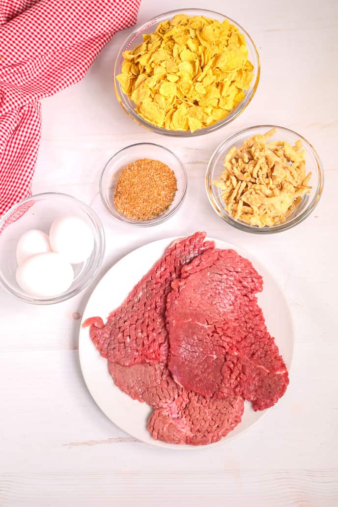 Finger Steaks on a white round plate with garlic, eggs and cornflakes with a red napkin in background.