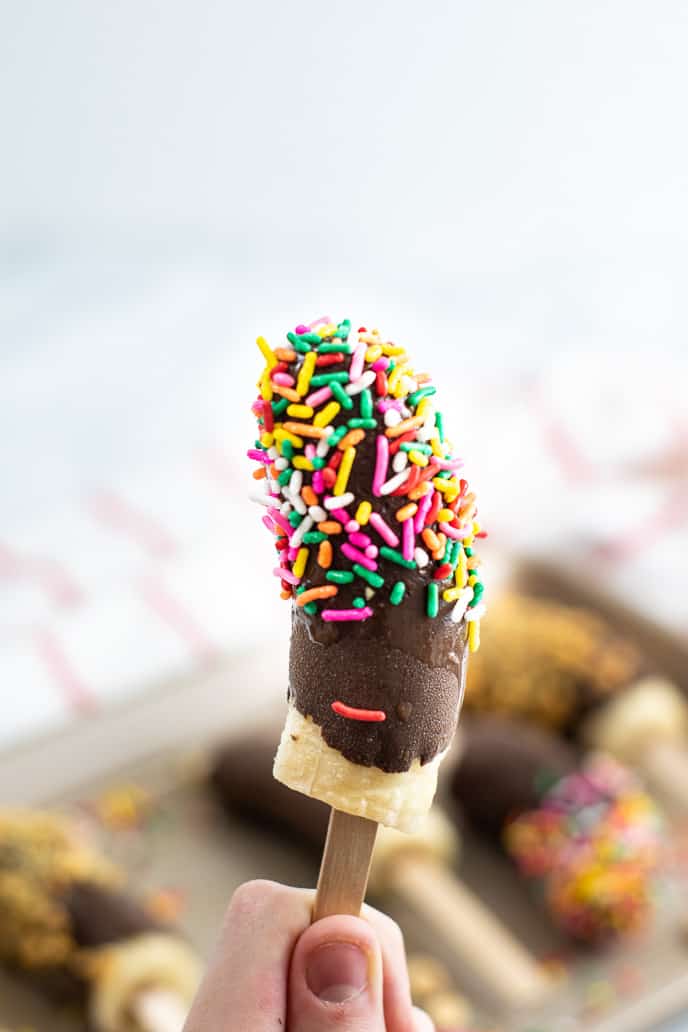 Hand holding a frozen banana with chocolate and multicolored sprinkles.
