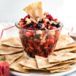 A clear glass bowl filled with a delightful fruit salsa, featuring diced apples, strawberries, and blueberries, surrounded by cinnamon-sugar coated tortilla chips on a white plate.
