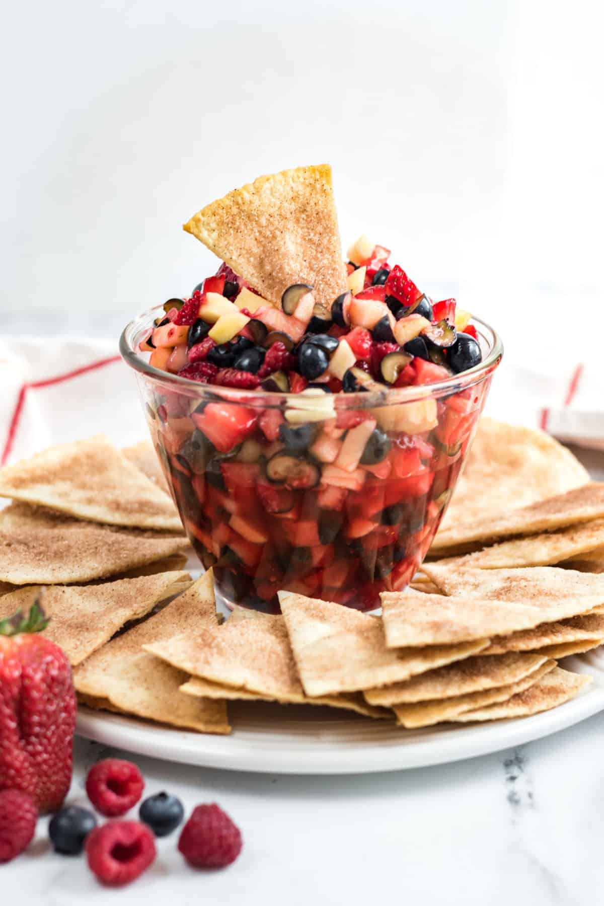 A clear glass bowl filled with a delightful fruit salsa, featuring diced apples, strawberries, and blueberries, surrounded by cinnamon-sugar coated tortilla chips on a white plate.
