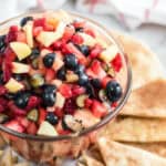 A glass bowl filled with a delicious fruit salsa of diced strawberries, blueberries, and apples is surrounded by triangular pieces of pita bread on a white surface.