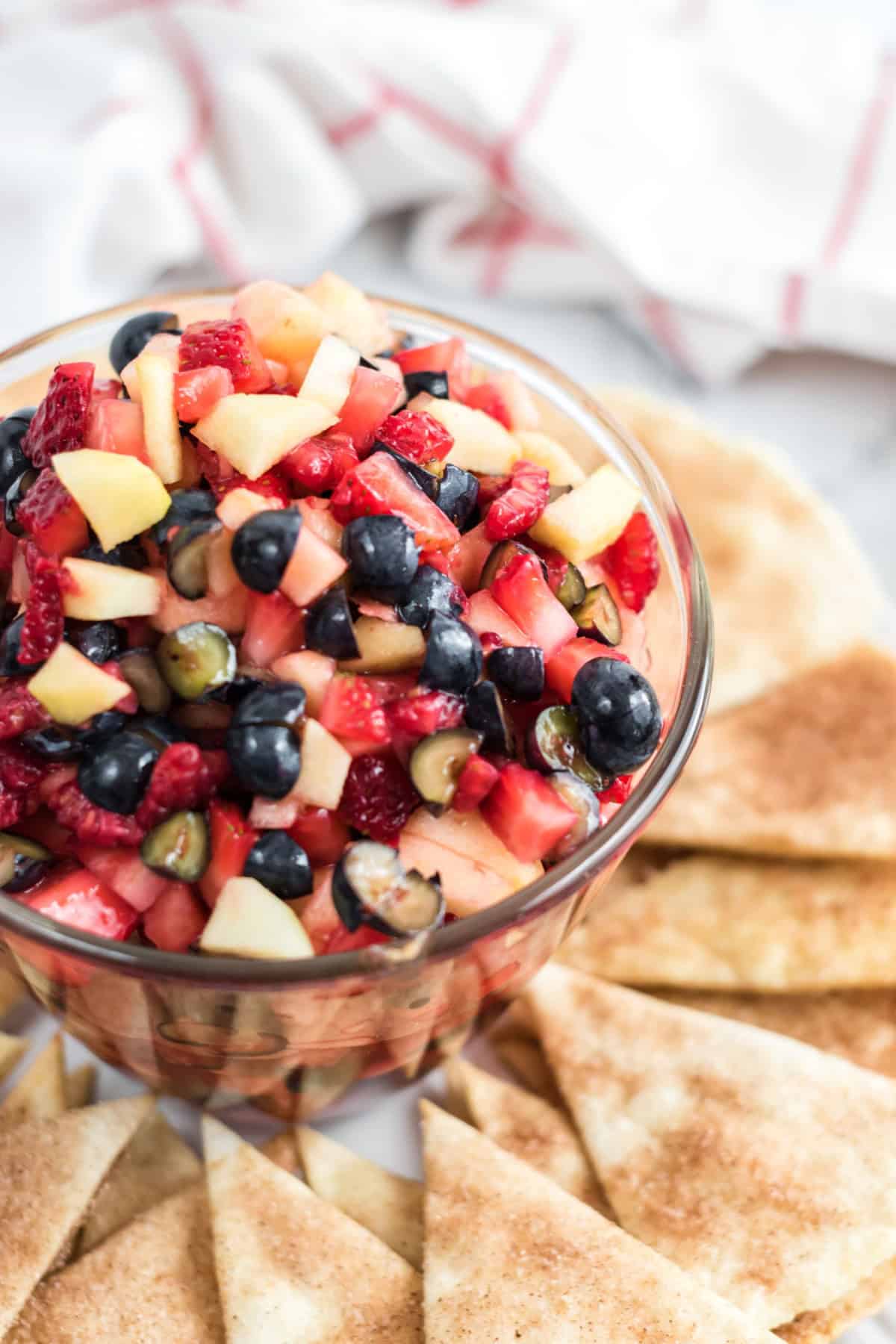 A glass bowl filled with a delicious fruit salsa of diced strawberries, blueberries, and apples is surrounded by triangular pieces of pita bread on a white surface.